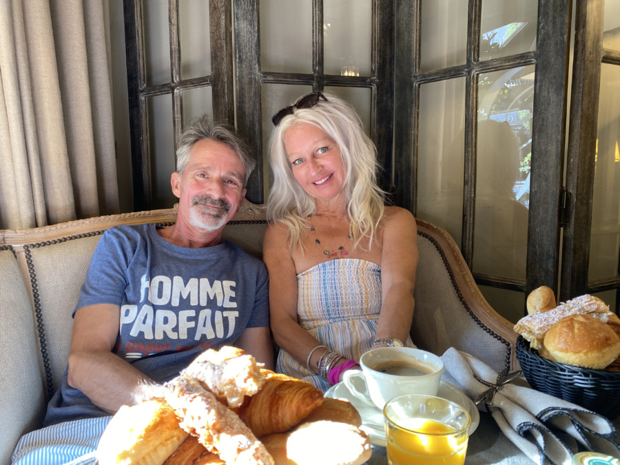 Christy and her husband sitting and smiling, with the coffee, orange juice, croissants on the table. 